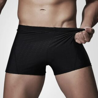 Maillot de bain noir boxer sur un homme blanc, qui tire sur son maillot pour en montrer l'élasticité.