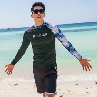 On voit un homme d'origine asiatique devant la plage qui porte un maillot de bain deux pièces composé d'un T-shirt manches longues et d'un short. Il a les bras écartés.