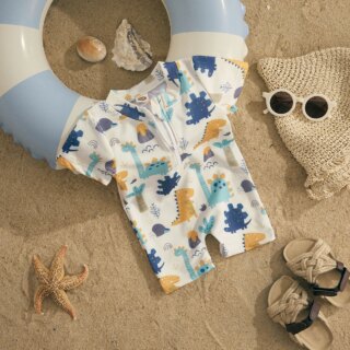 Exposé sur le sable, on voit un maillot de bain combinaison, blanc avec un imprimé dinosaure. Il est conçu pour les bébés et disposé sur une bouée bleue et blanche, près de lunettes de soleil et d'une étoile de mer.