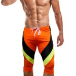 On voit un homme qui porte un long maillot de bain en dessous du genou. Il est orange et noir.