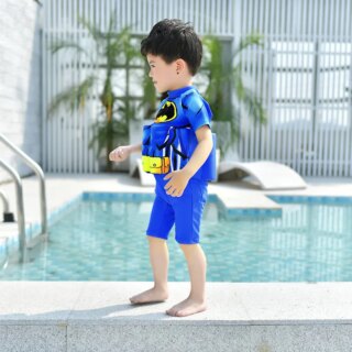 On voit un petit garçon qui se promène sur le bord d'une piscine d'hôtel. Il porte un maillot de bain bouée bleu à l'effigie de Batman.