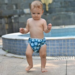 On voit un bébé devant une piscine carrelée qui porte un maillot de bain, une couche de bain bleu-vert avec des baleines. Le bébé est brun et doit avoir à peine plus d'un an. Il marche.