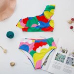 Photo d'un maillot de bain deux pièces multicolore pour fille adolescente composé d'une culotte haute et d'un haut asymétrique à volant sur une table avec un magazine et des accessoires