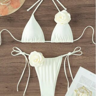 Photo d'un maillot de bain blanc ficelle avec une rose en 3D en haut du sein gauche et en bas du bikini posé sur un fond bois avec chapeau et lunettes dans le coin gauche