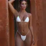 Photo représentant une femme debout le bras droit levé coude plié derrière la tête portant un maillot de bain deux pièces ficelle string blanc sur un fond marron